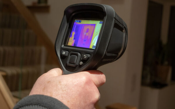 A male surveyor using a handheld infrared thermal camera for a heat loss survey, to inspect the interior of a domestic home, looking for heat anomalies. The camera viewfinder shows a coloured thermal image with varying temperature ranges.