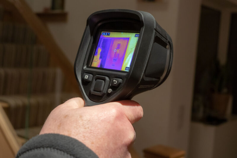 A male surveyor using a handheld infrared thermal camera for a heat loss survey, to inspect the interior of a domestic home, looking for heat anomalies. The camera viewfinder shows a coloured thermal image with varying temperature ranges.