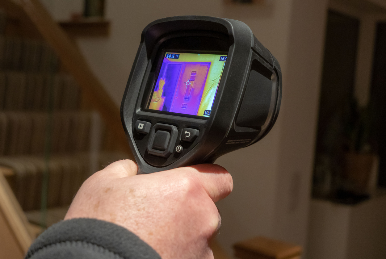 A male surveyor using a handheld infrared thermal camera for a heat loss survey, to inspect the interior of a domestic home, looking for heat anomalies. The camera viewfinder shows a coloured thermal image with varying temperature ranges.