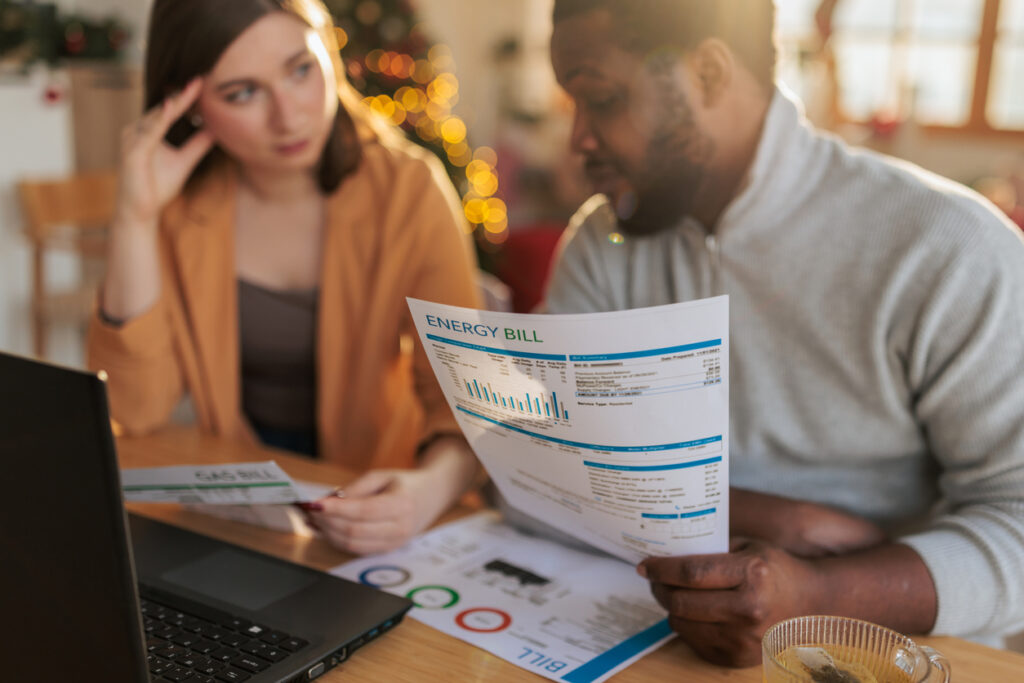 A couple sitting at a table at home, examining an energy bill while using a laptop. The man holds a printed utility statement with charts and usage details, while the woman studies additional documents beside him.