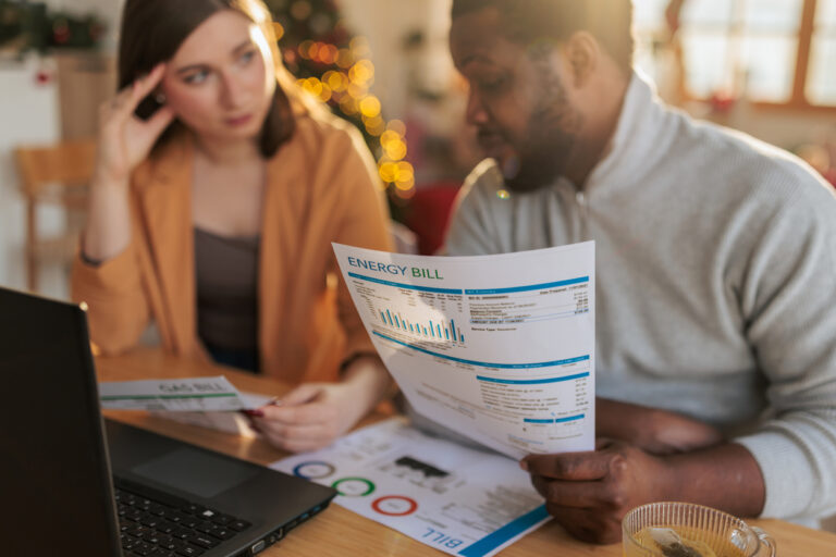 A couple sitting at a table at home, examining an energy bill while using a laptop. The man holds a printed utility statement with charts and usage details, while the woman studies additional documents beside him.