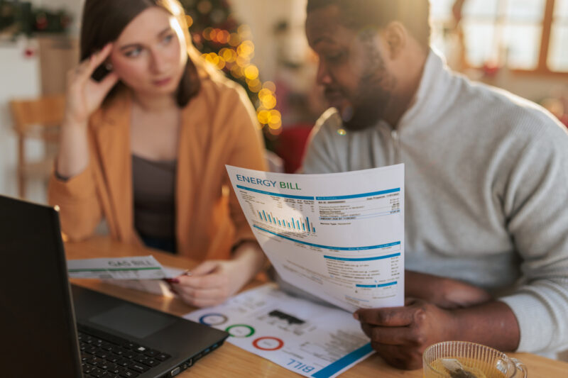 A couple sitting at a table at home, examining an energy bill while using a laptop. The man holds a printed utility statement with charts and usage details, while the woman studies additional documents beside him.