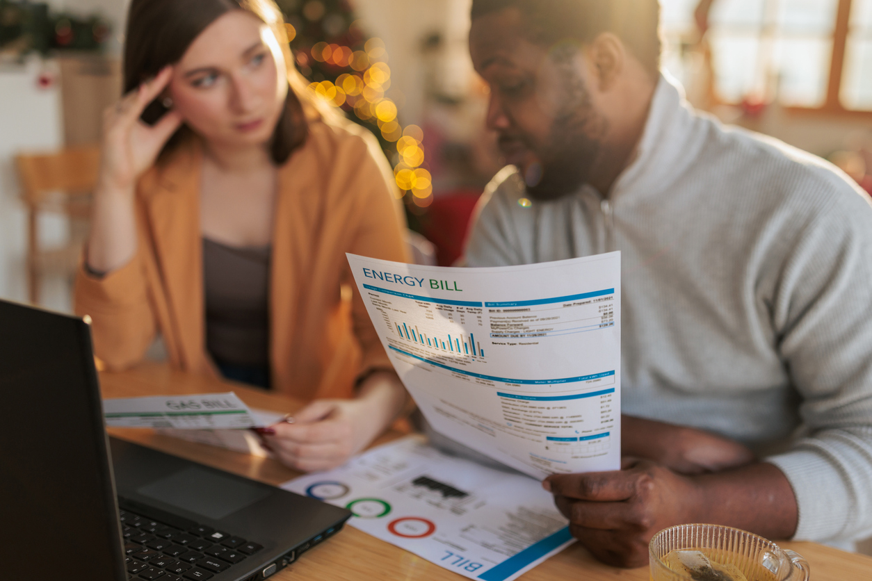 A couple sitting at a table at home, examining an energy bill while using a laptop. The man holds a printed utility statement with charts and usage details, while the woman studies additional documents beside him.